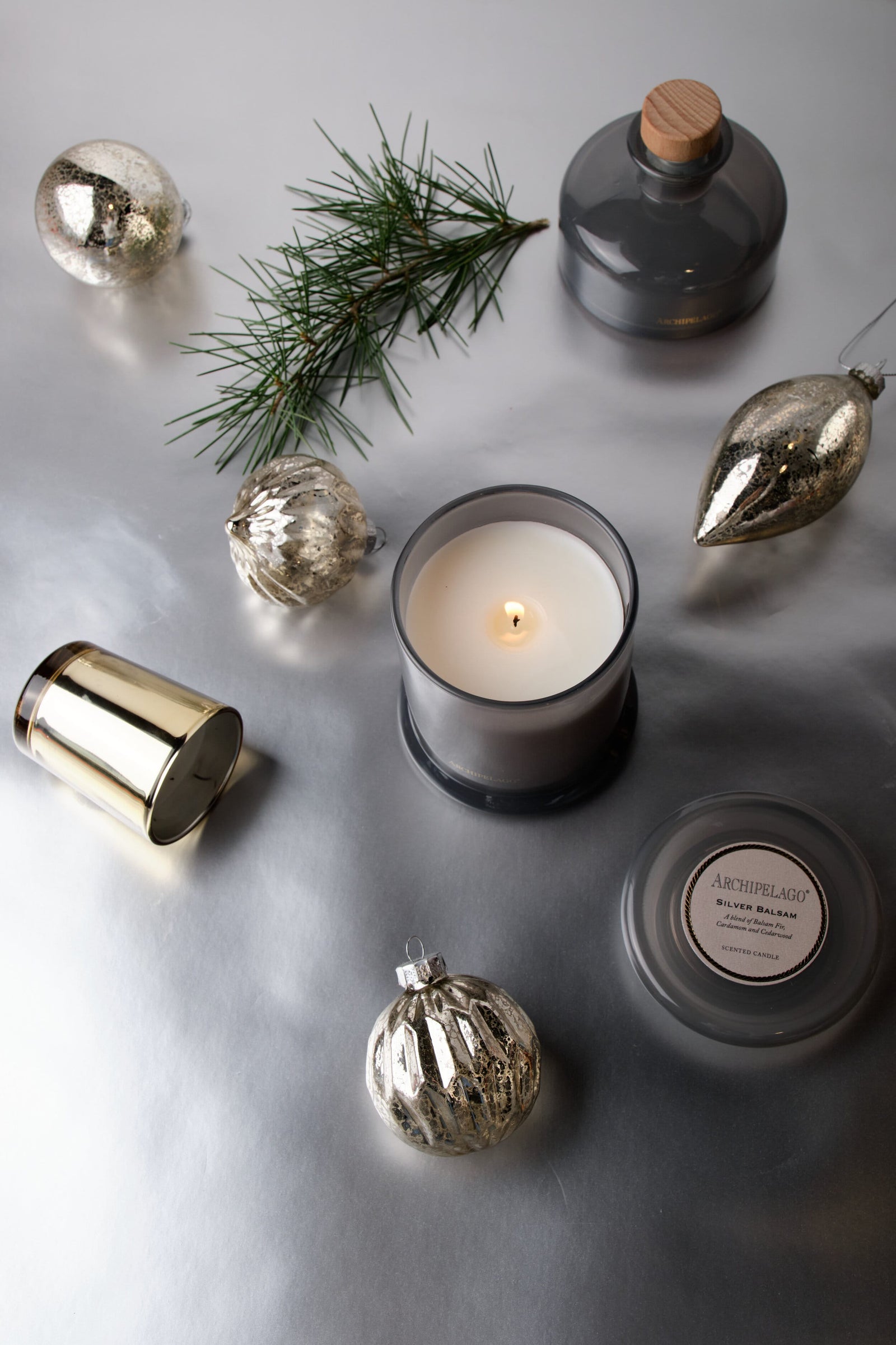 A Silver Balsam Petite Candle in a grey glass jar surrounded by Christmas ornaments, a pine sprig, and a matching candle lid on a silver surface. - image 2