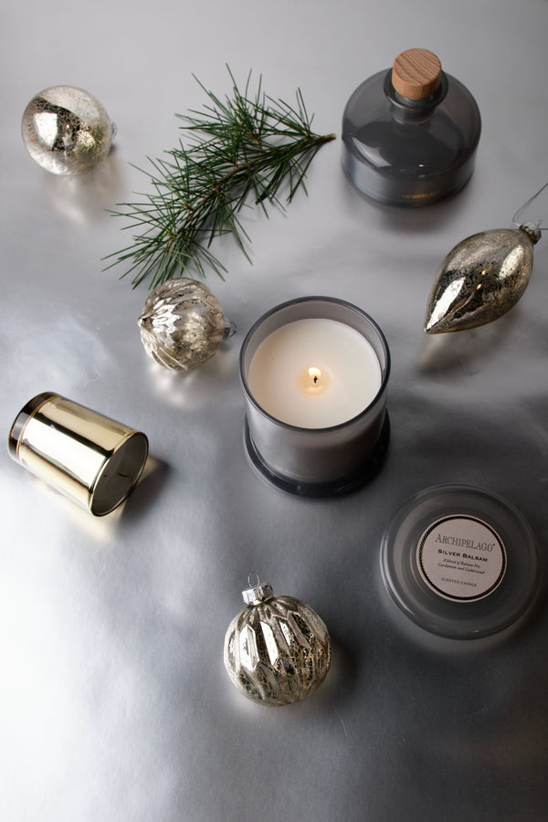 A Silver Balsam Petite Candle in a grey glass jar surrounded by Christmas ornaments, a pine sprig, and a matching candle lid on a silver surface.