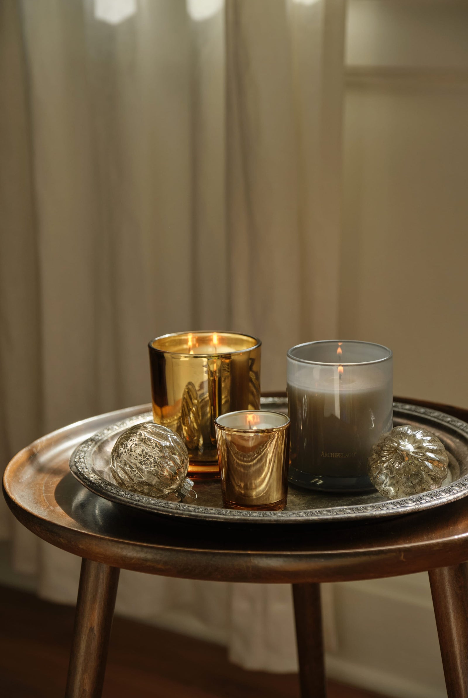 Three Silver Balsam Petite candles in gold glass vessels and two glass ornaments arranged on a decorative metal tray on a wooden side table. - image 3