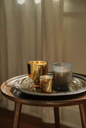 Three Silver Balsam Petite candles in gold glass vessels and two glass ornaments arranged on a decorative metal tray on a wooden side table. - image 3