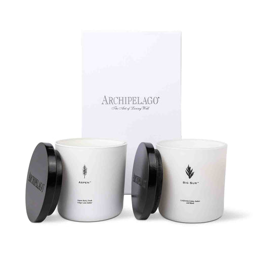 Two luxe candles, Aspen and Big Sur, made with coconut wax and black lids, sit in front of The Mountains Luxe Candle Gift Set box.