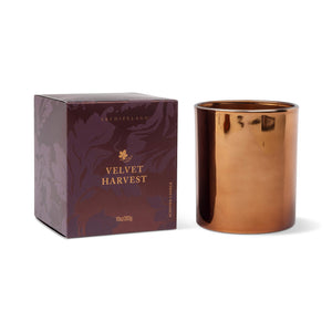 The Velvet Harvest Boxed Candle features a brown glass jar with a metallic, reflective finish and is presented alongside a purple box adorned with gold text, leaf designs, and the Archipelago branding. With its sandalwood scent evoking a cozy fall fragrance, this candle makes an ideal gift. - image 1