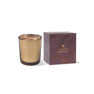 A Velvet Harvest Boxed Votive Candle, featuring a gold-colored candle in a glass holder, is displayed beside its purple box adorned with decorative leaf patterns and “Archipelago” branding in gold text. - image 1