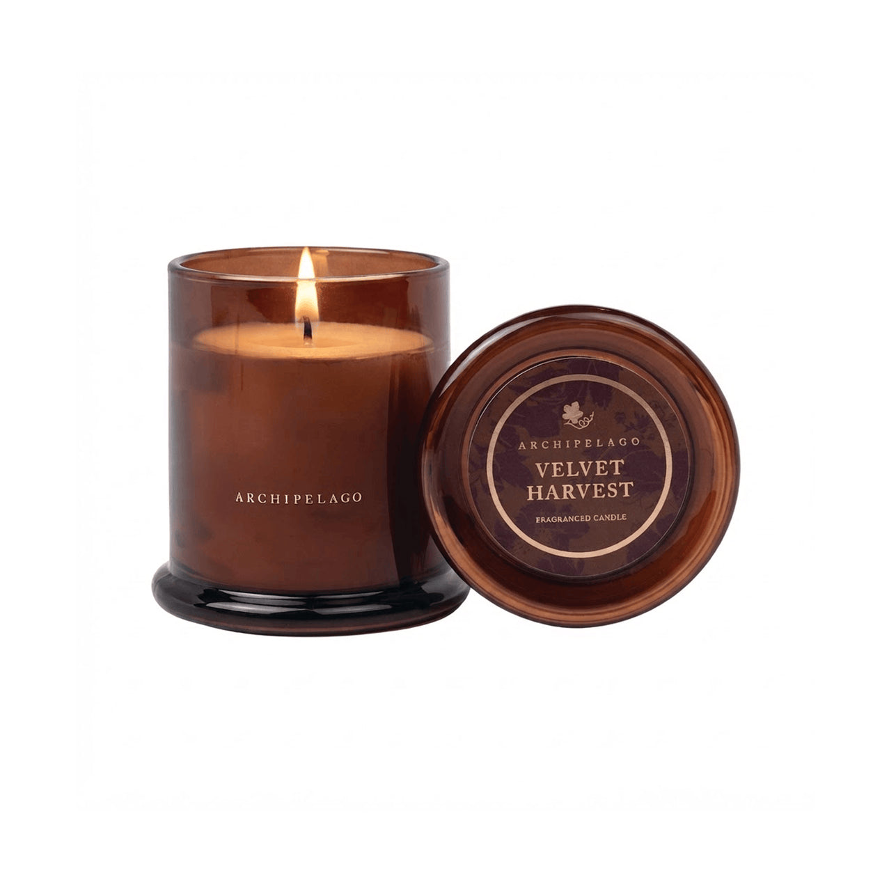 A brown glass Velvet Harvest Jar Candle burns beside its lid, which reads “Archipelago Velvet Harvest Fragranced Candle,” on a white background, evoking warm fall fragrances with subtle notes of sandalwood. - image 1