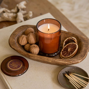 A lit Velvet Harvest Jar Candle in a brown glass holder sits in a wooden tray alongside whole walnuts and dried orange slices. Nearby, a matching tin lid and a small plate with matches complete the scene, creating a cozy, rustic atmosphere perfect for savoring seasonal fall fragrances. - image 3