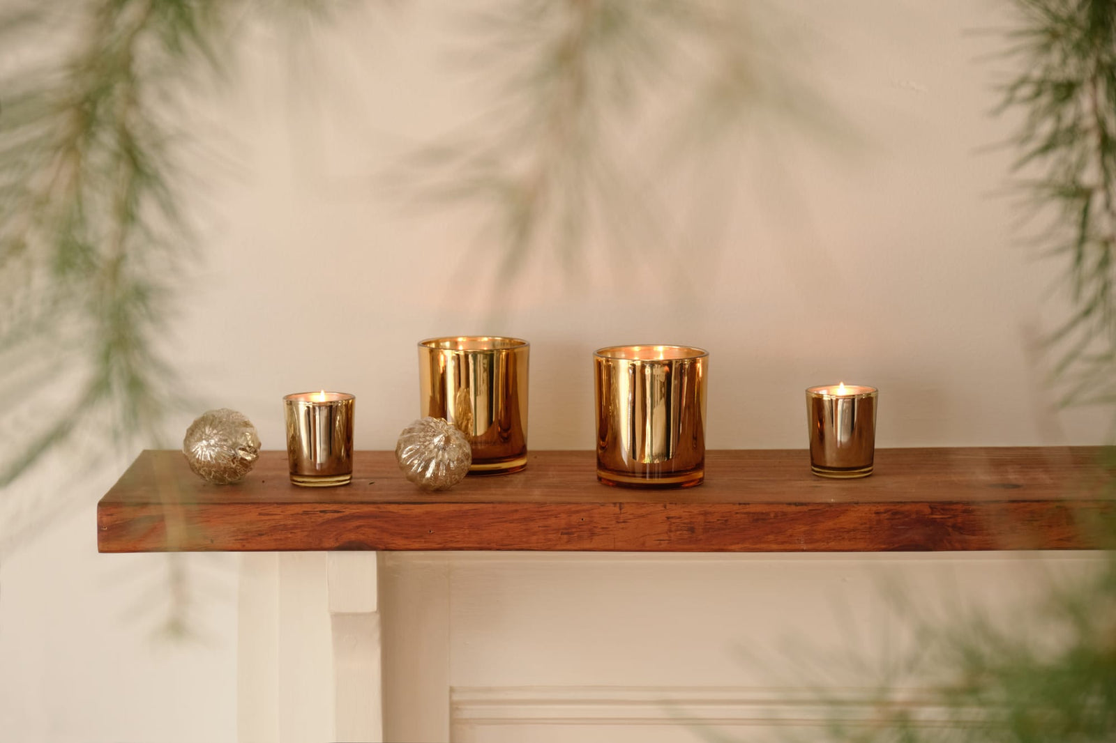 Four Winter Frost Boxed Petite Candle candles in gold glass votives and two decorative metallic spheres arranged on a wooden mantel. - image 3