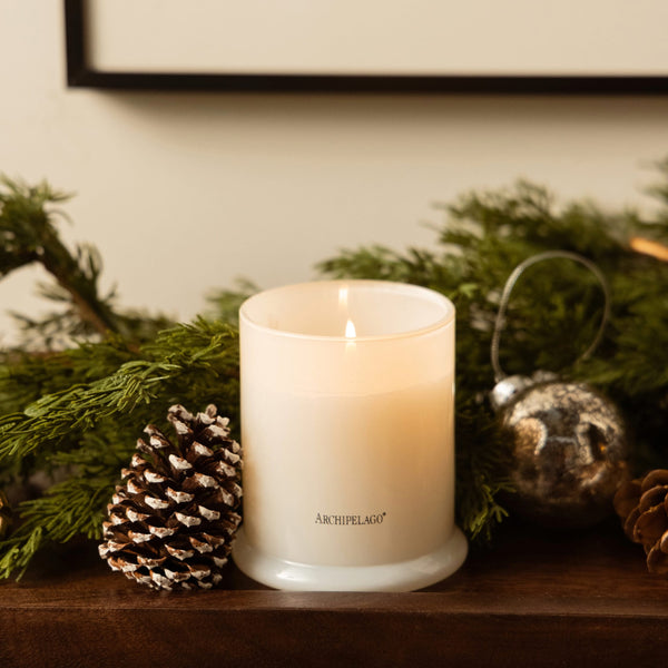 A Winter Frost Jar Candle in a frosted glass jar, centered in front of evergreen branches, a pinecone, and a silver ornament on a wooden surface.