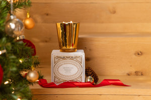 A Winter Frost Mantel Candle in a gold glass jar sits atop its decorative white box on a wooden surface beside a Christmas tree. - image 3