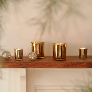 Four gold metallic Winter Frost Petite candles in various sizes, some lit, arranged on a wooden mantelpiece with two spherical ornaments. - image 2