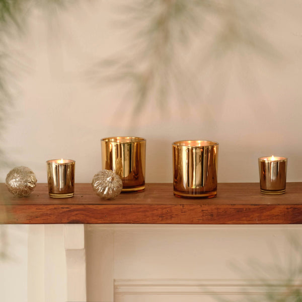 Four gold metallic Winter Frost Petite candles in various sizes, some lit, arranged on a wooden mantelpiece with two spherical ornaments.