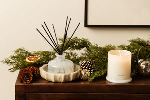 A Winter Frost Reed Diffuser in a frosted glass bottle sits in a stone bowl next to a lit white candle, surrounded by evergreen branches, pinecones, and holiday ornaments on a wooden surface. - image 3